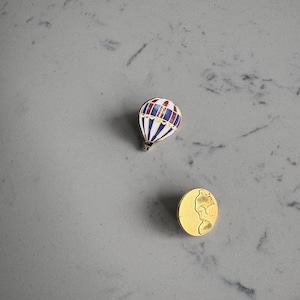 May include: A miniature hot air balloon pin with a red, white, and blue striped design. Next to it is a gold-colored round pin with a profile of a person. Both pins are on a gray surface.