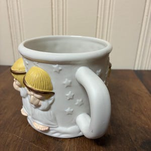 May include: White ceramic mug with a detailed relief design of figures wearing yellow hats and robes. The mug features a white handle and star-shaped accents. The mug is sitting on a wooden surface.