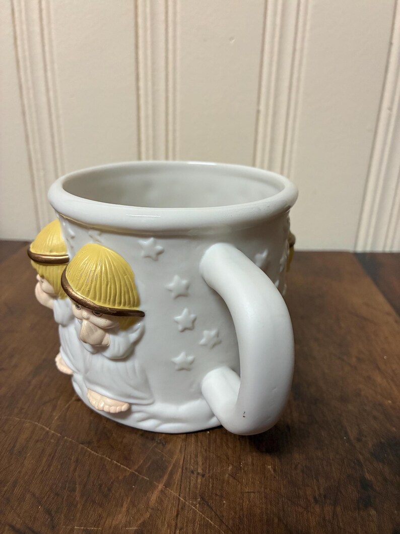 May include: White ceramic mug with a detailed relief design of figures wearing yellow hats and robes. The mug features a white handle and star-shaped accents. The mug is sitting on a wooden surface.