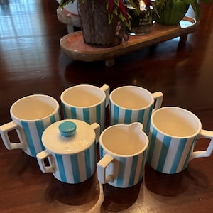 May include: A set of six ceramic mugs, a creamer, and a sugar bowl with a lid. The mugs and creamer have a white base with vertical turquoise stripes. The sugar bowl has a turquoise lid. The set is on a dark wooden surface.