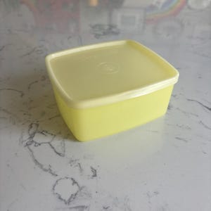 May include: A square, pale yellow plastic food storage container with a matching lid. The lid features a subtle, embossed circular design in the center and a slightly raised edge, providing a secure seal for the base of the container.