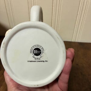 May include: A white ceramic mug with a curved handle. The base of the mug features the text "PRODUCED FOR HOUSTON HARVEST GIFT PRODUCTS LLC" and "&copy; Hallmark Licensing, Inc.". The mug is made in China.