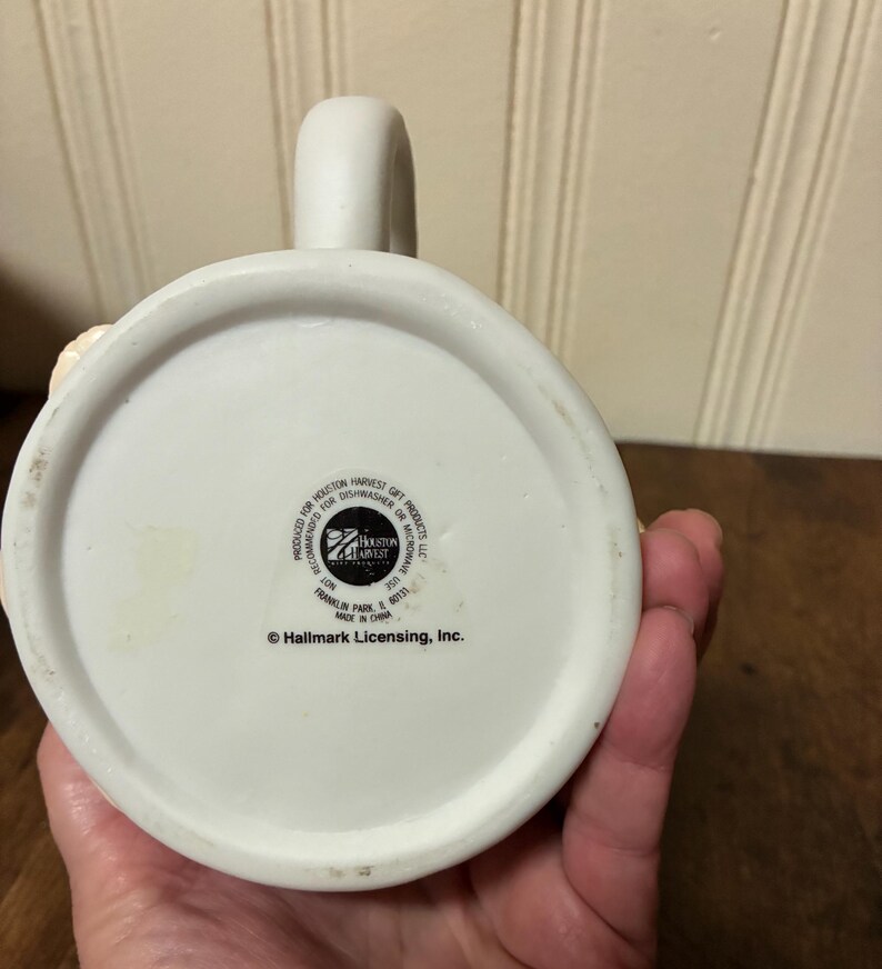 May include: A white ceramic mug with a curved handle. The base of the mug features the text "PRODUCED FOR HOUSTON HARVEST GIFT PRODUCTS LLC" and "&copy; Hallmark Licensing, Inc.". The mug is made in China.