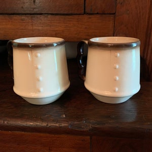 May include: Two white ceramic mugs with brown rims and three raised dots on the front. The mugs have brown handles.
