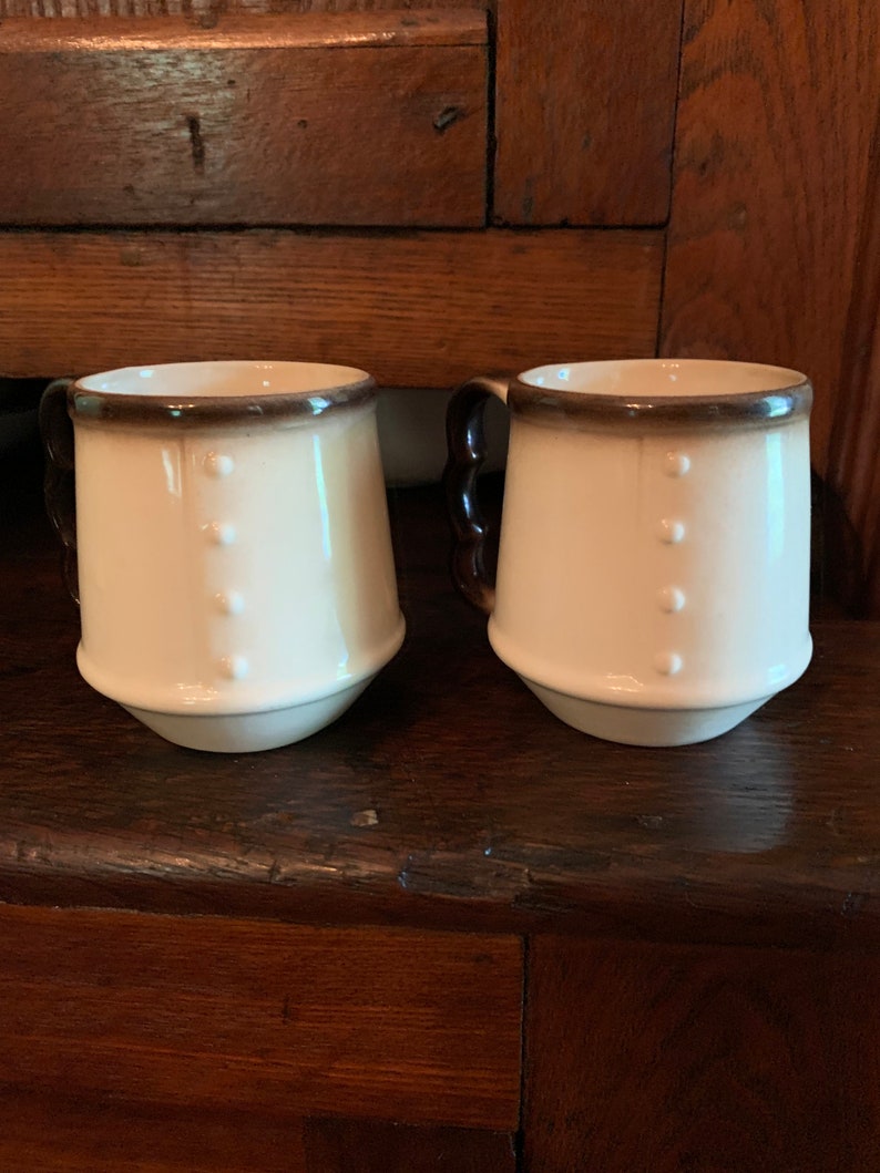 May include: Two white ceramic mugs with brown rims and three raised dots on the front. The mugs have brown handles.