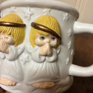 May include: White ceramic mug with embossed angels, yellow hair, and halos. The angels are depicted in prayer, hands clasped. The mug has a handle and is decorated with star-shaped accents.