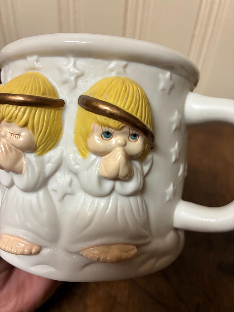 May include: White ceramic mug with embossed angels, yellow hair, and halos. The angels are depicted in prayer, hands clasped. The mug has a handle and is decorated with star-shaped accents.