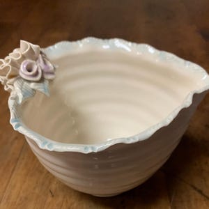 May include: A white ceramic bowl with a ruffled edge and a delicate floral design. The bowl is hand-crafted and has a unique, organic shape.