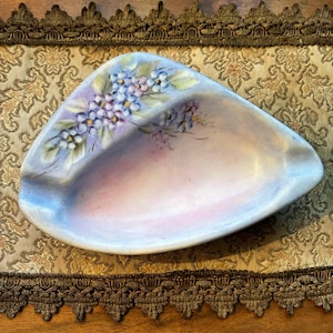 May include: A hand-painted, ceramic ashtray with a unique, asymmetrical shape. The ashtray features a soft, pastel color palette of blue, pink, and lavender. Delicate floral designs in blue, purple, and green adorn the rim, adding a touch of elegance.