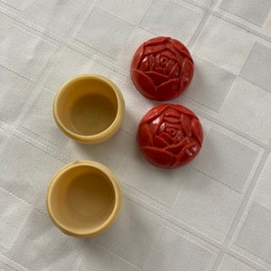May include: Two small, round, red boxes with a rose design on the top and two small, round, cream-colored boxes.