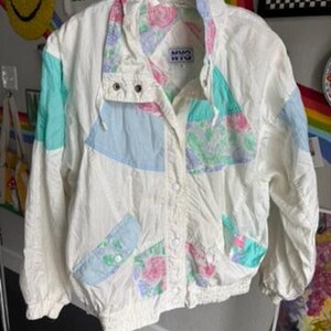 May include: A white windbreaker jacket with a floral print and a blue and green color scheme. The jacket has a zipper closure and a stand-up collar. The brand name "Nino Rita" is printed on the front of the jacket.