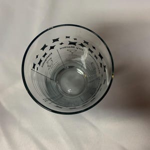 Puede incluir: Un vaso medidor de vidrio transparente con marcas negras para hacer un martini seco. Las marcas incluyen medidas para ginebra, vermut y aceitunas.