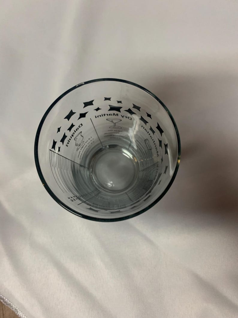 Puede incluir: Un vaso medidor de vidrio transparente con marcas negras para hacer un martini seco. Las marcas incluyen medidas para ginebra, vermut y aceitunas.