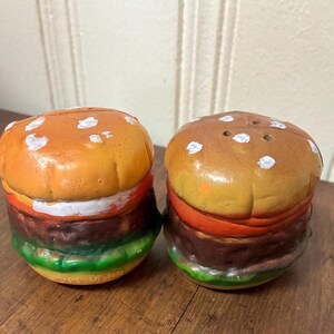 May include: Two ceramic salt and pepper shakers shaped like hamburgers. The shakers have orange, brown, red, and green painted details. The top bun is orange with white dots, and the bottom bun is green.
