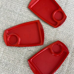 May include: Three red plastic trays with a small circular indentation on each. The trays are on a woven beige surface.