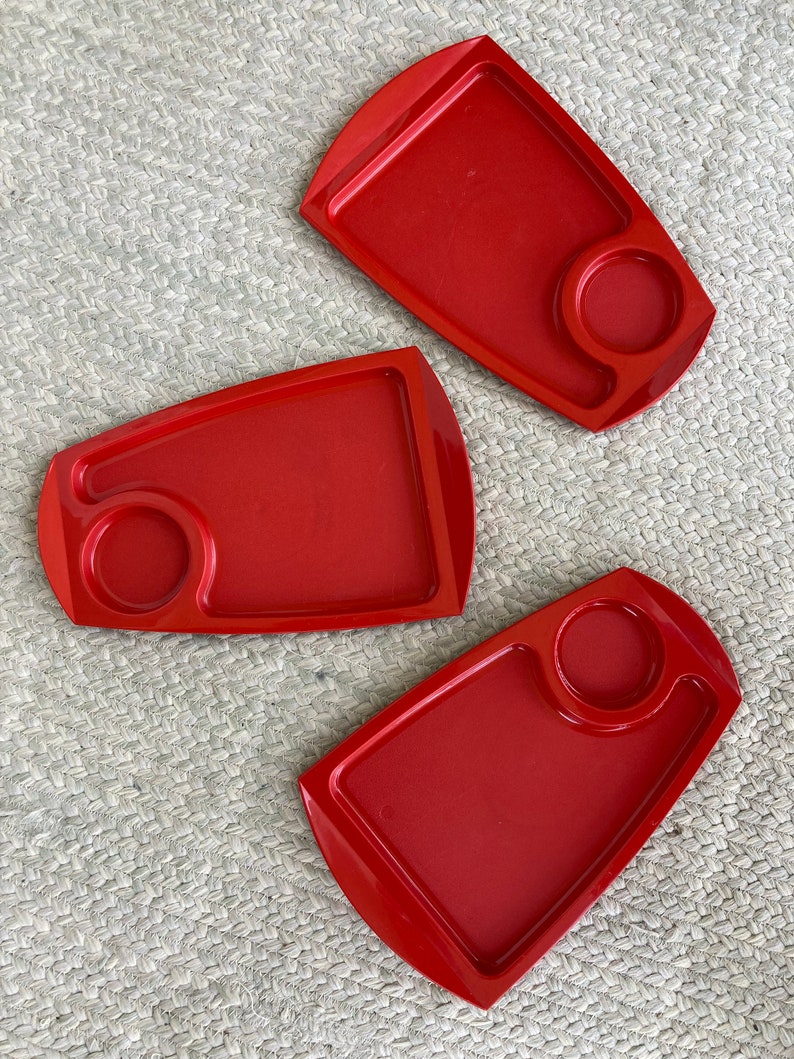 May include: Three red plastic trays with a small circular indentation on each. The trays are on a woven beige surface.