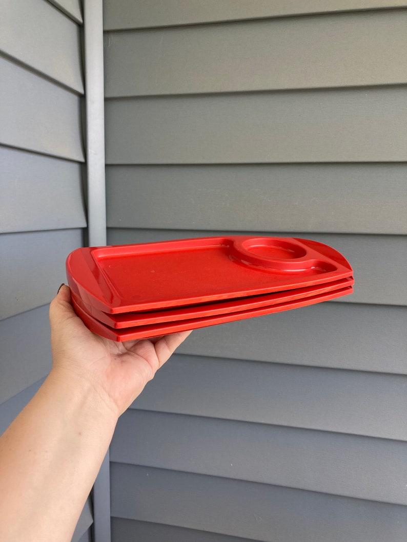 May include: Three red plastic trays with a small indented circle on the top tray. The trays are stacked on top of each other.