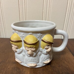 May include: White ceramic mug featuring three angel figures with yellow hair and gold halos. The angels are in prayer, with hands clasped. The mug has a white handle and is decorated with star shapes.