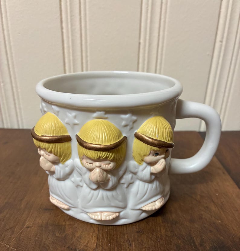 May include: White ceramic mug featuring three angel figures with yellow hair and gold halos. The angels are in prayer, with hands clasped. The mug has a white handle and is decorated with star shapes.