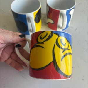 K&ouml;nnte beinhalten: Drei Keramikbecher mit farbenfrohen abstrakten Designs. Ein Becher wird von einer Hand gehalten. Die Becher haben einen wei&szlig;en Griff und Innenraum mit Au&szlig;endesigns in Rot, Gelb, Blau und Schwarz. Die Becher sind wahrscheinlich f&uuml;r Kaffee oder Tee.