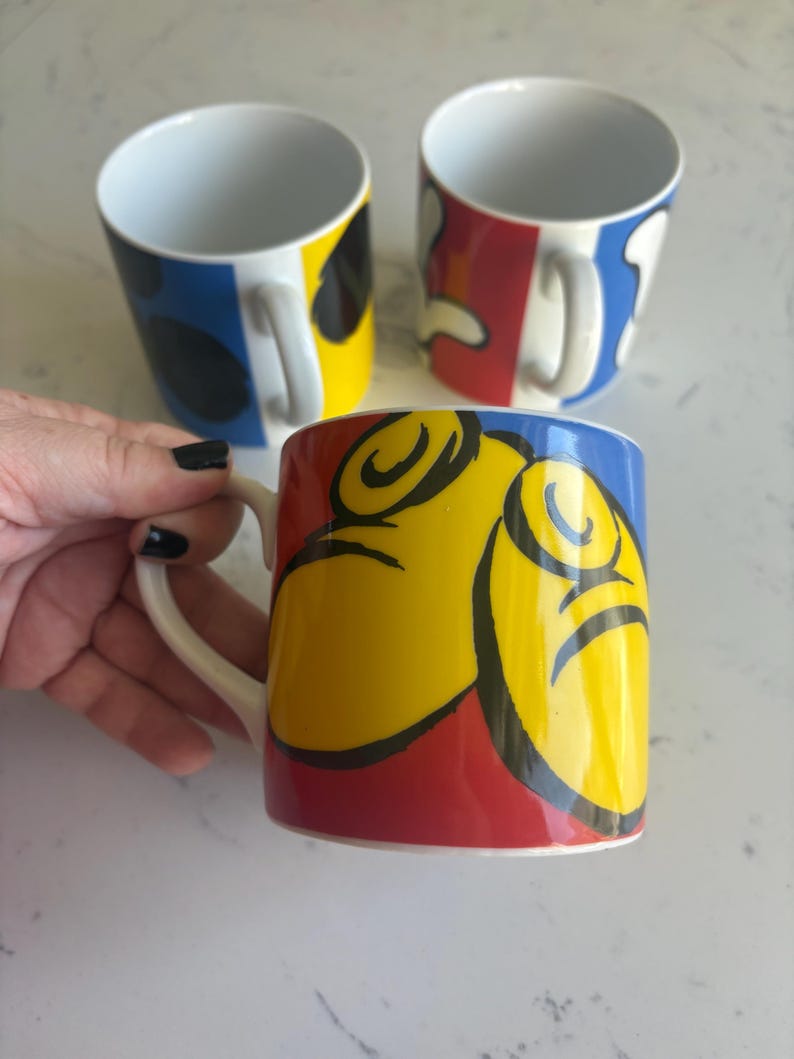 K&ouml;nnte beinhalten: Drei Keramikbecher mit farbenfrohen abstrakten Designs. Ein Becher wird von einer Hand gehalten. Die Becher haben einen wei&szlig;en Griff und Innenraum mit Au&szlig;endesigns in Rot, Gelb, Blau und Schwarz. Die Becher sind wahrscheinlich f&uuml;r Kaffee oder Tee.
