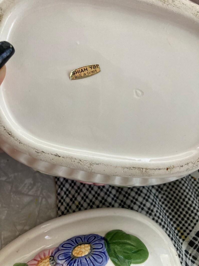 May include: White ceramic lidded dish with a floral design. The lid has a gold sticker that reads "Shiah Yihi Made in Taiwan."