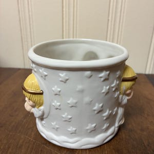 May include: White ceramic planter with embossed star pattern and two angel figures. The planter has a round shape with a wide opening and is suitable for small plants or decorative use. The angels have blonde hair and gold halos.