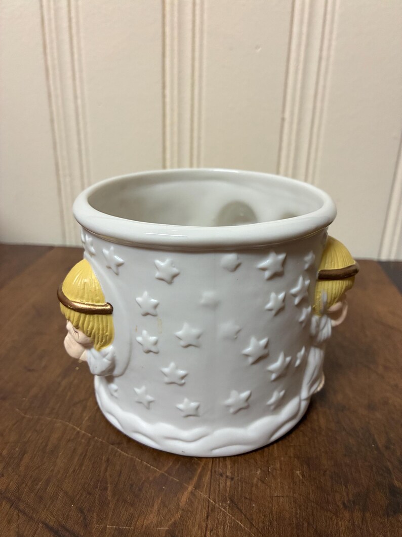 May include: White ceramic planter with embossed star pattern and two angel figures. The planter has a round shape with a wide opening and is suitable for small plants or decorative use. The angels have blonde hair and gold halos.
