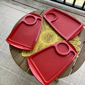 May include: Three red plastic trays with a small circular indentation in the center of each tray. The trays are on a wooden table with a yellow doily.
