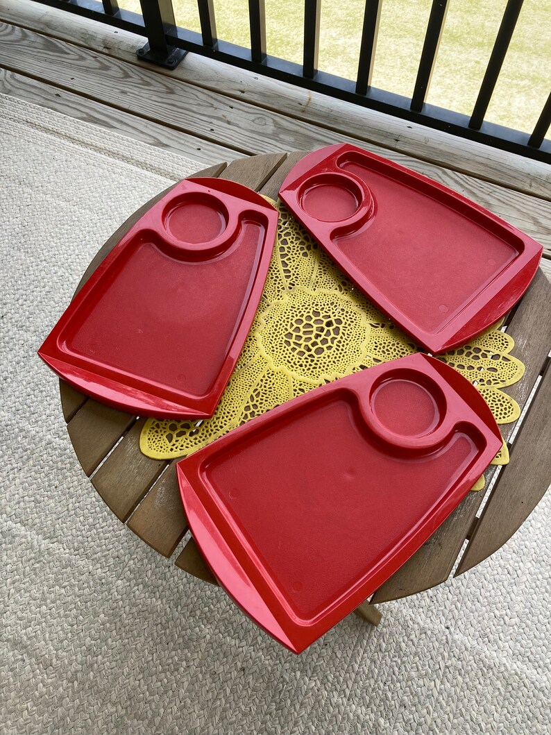 May include: Three red plastic trays with a small circular indentation in the center of each tray. The trays are on a wooden table with a yellow doily.