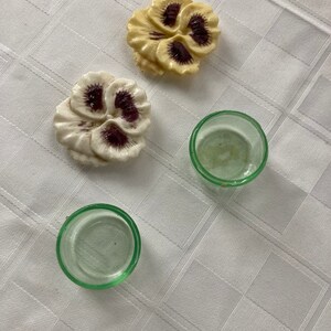 May include: Two ceramic pansy flower decorations in white and yellow with purple centers, and two clear glass candle holders with a green tint.