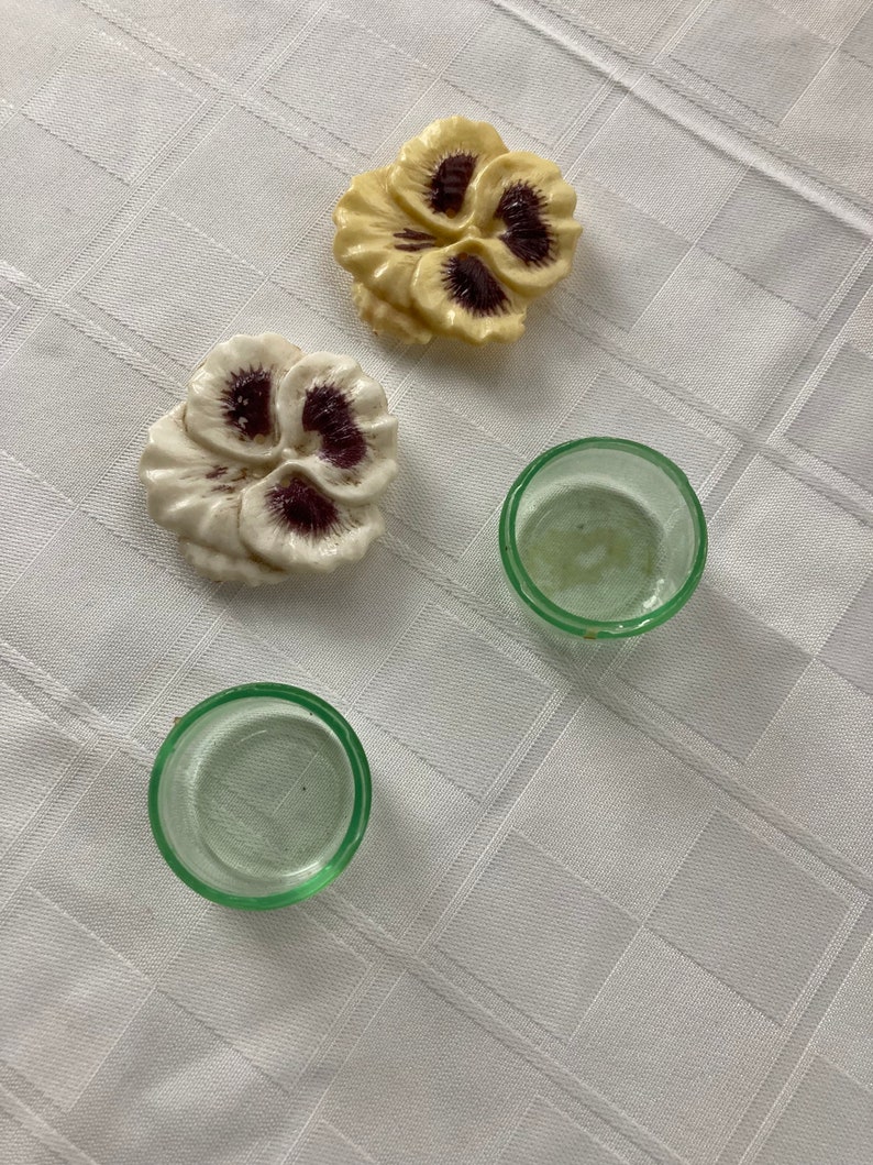 May include: Two ceramic pansy flower decorations in white and yellow with purple centers, and two clear glass candle holders with a green tint.