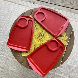 May include: Three red plastic trays with a small circular indentation in the center of each tray. The trays are on a wooden table with a yellow crocheted doily.