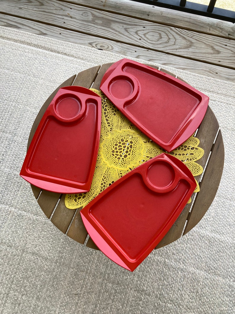 May include: Three red plastic trays with a small circular indentation in the center of each tray. The trays are on a wooden table with a yellow crocheted doily.