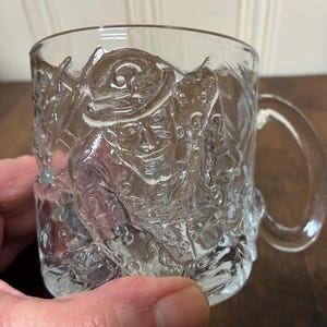 May include: Clear glass mug with a detailed embossed design featuring a figure in a hat and ornate patterns. The mug has a curved handle and is held by a hand. The mug is made of clear glass.