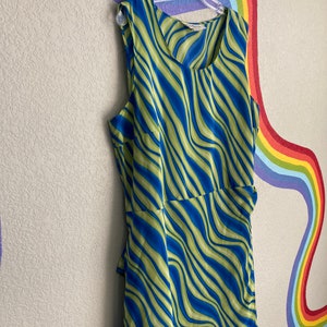 May include: A sleeveless dress with a green and blue wavy pattern. The dress is hanging on a hanger against a wall with a rainbow painted on it.