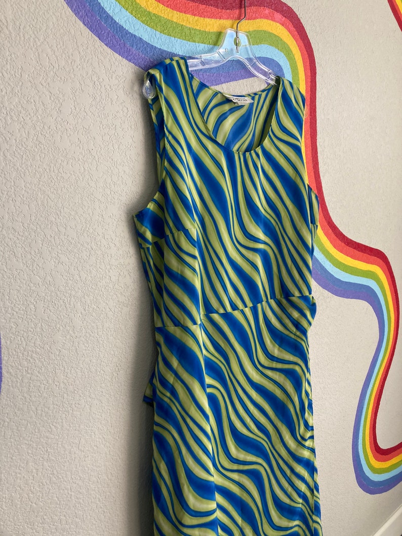 May include: A sleeveless dress with a green and blue wavy pattern. The dress is hanging on a hanger against a wall with a rainbow painted on it.