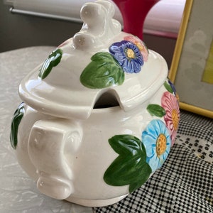 May include: White ceramic serving dish with a lid. The dish is decorated with green leaves and colorful flowers. The lid has a knob in the shape of a flower.