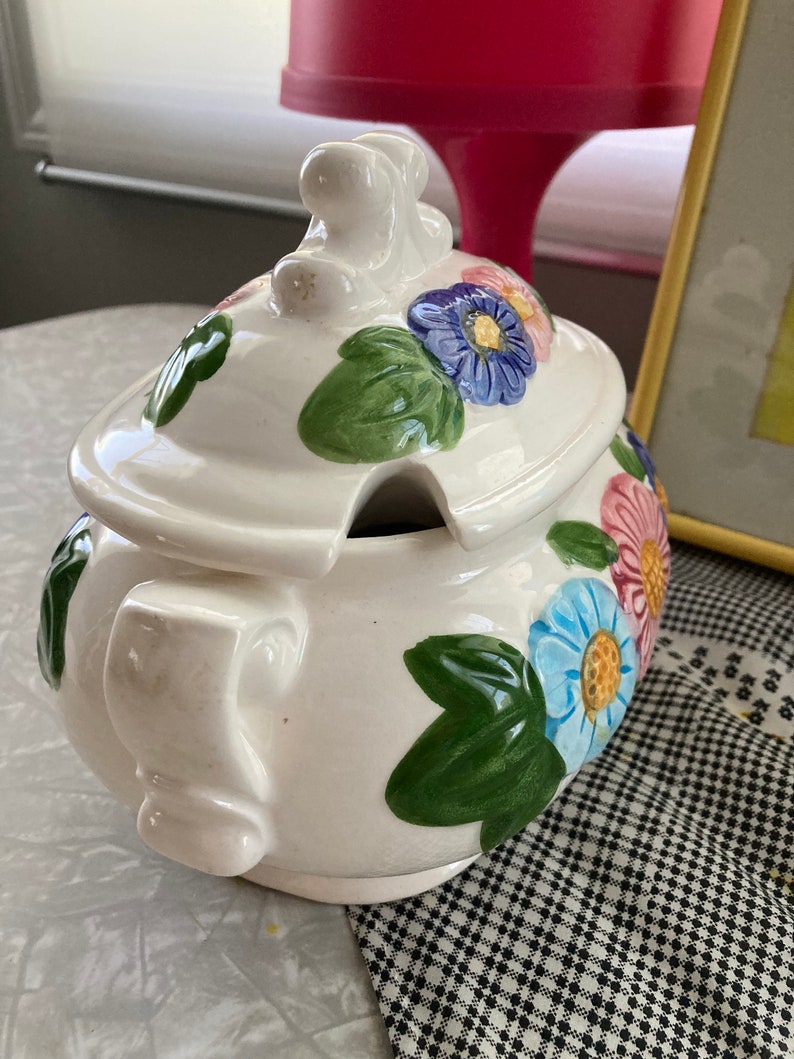 May include: White ceramic serving dish with a lid. The dish is decorated with green leaves and colorful flowers. The lid has a knob in the shape of a flower.