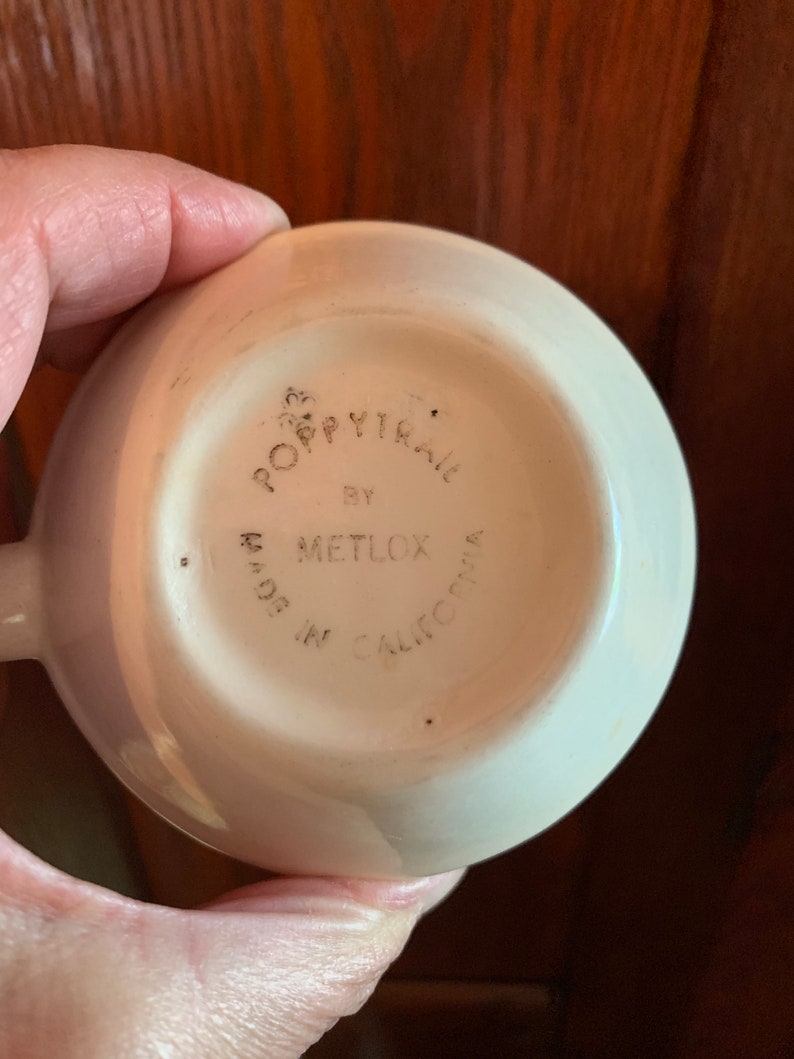 May include: The bottom of a white ceramic cup with the words "Poppytrail by Metlox Made in California" printed on it.