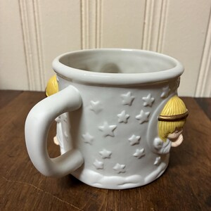 May include: White ceramic mug featuring embossed stars and two angel figures. The mug has a curved handle and a slightly flared rim. The angels have yellow hair and are in relief. A charming and decorative item.