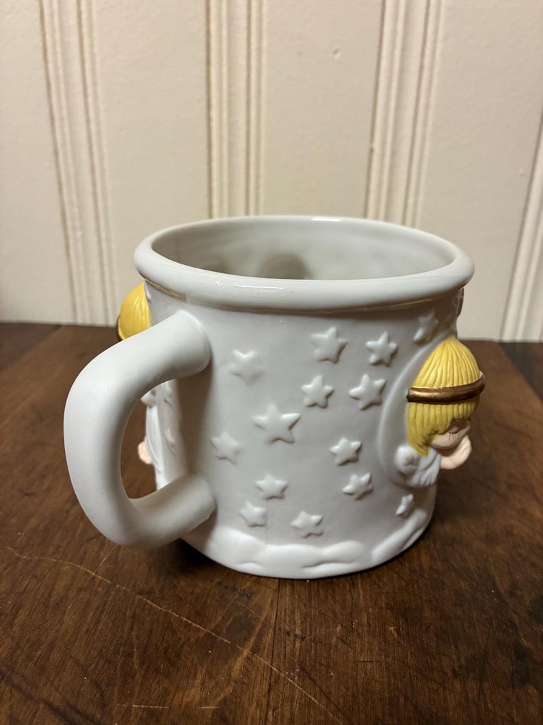 May include: White ceramic mug featuring embossed stars and two angel figures. The mug has a curved handle and a slightly flared rim. The angels have yellow hair and are in relief. A charming and decorative item.