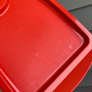 May include: A bright red plastic container lid with a slightly rounded edge. The lid has a smooth surface with a few scratches.