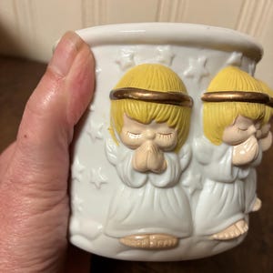 May include: White ceramic candle holder featuring embossed praying angel figures. The angels have yellow hair, gold halos, and white robes. The candle holder is decorated with star shapes. A decorative item for home decor.