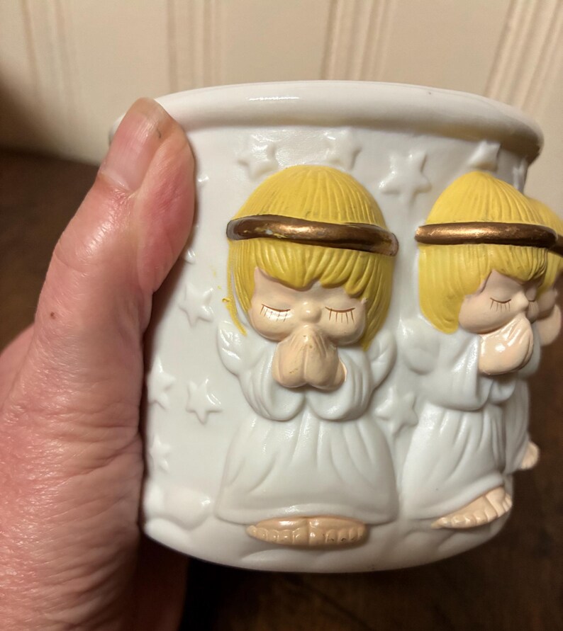 May include: White ceramic candle holder featuring embossed praying angel figures. The angels have yellow hair, gold halos, and white robes. The candle holder is decorated with star shapes. A decorative item for home decor.