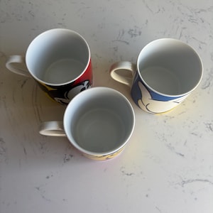 K&ouml;nnte beinhalten: Drei wei&szlig;e Keramikbecher mit bunten Cartoon-Charakter-Designs. Ein Becher hat ein rot-schwarzes Design, ein anderer ein blau-gelbes Design und der dritte ein gelb-rotes Design. Jeder Becher hat einen wei&szlig;en Henkel.