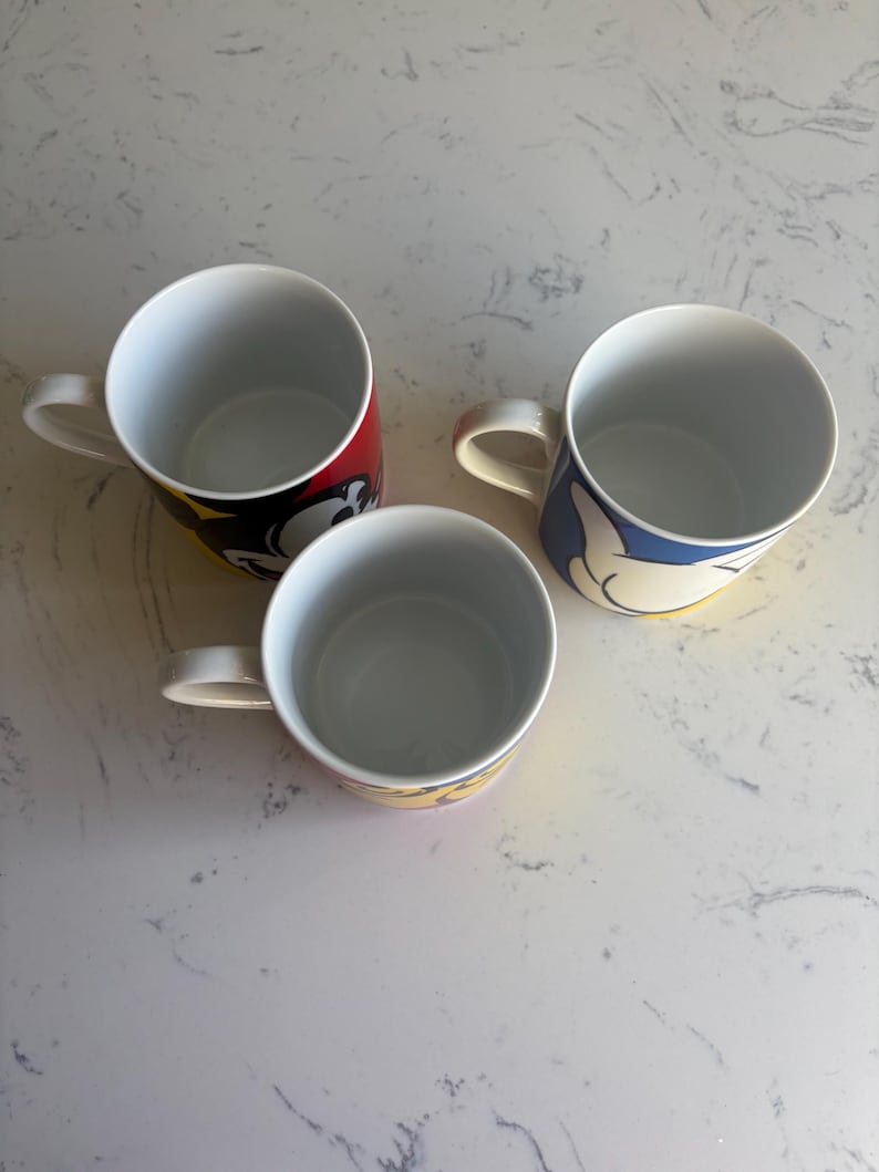 K&ouml;nnte beinhalten: Drei wei&szlig;e Keramikbecher mit bunten Cartoon-Charakter-Designs. Ein Becher hat ein rot-schwarzes Design, ein anderer ein blau-gelbes Design und der dritte ein gelb-rotes Design. Jeder Becher hat einen wei&szlig;en Henkel.