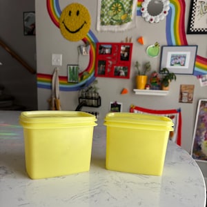 May include: Two square, yellow plastic containers with lids sit on a white surface. The containers are side-by-side, and the lids are flush with the container's edges. The background features a wall decorated with art and a rainbow.