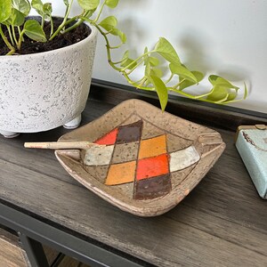 May include: A square ceramic ashtray with a geometric pattern of brown, orange, and tan squares. A lit cigarette rests in the ashtray's groove. The ashtray sits on a dark wooden surface, with a potted plant in the background.