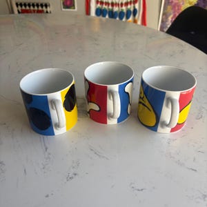K&ouml;nnte beinhalten: Drei Keramikbecher mit farbenfrohen abstrakten Designs. Jeder Becher hat einen wei&szlig;en Henkel und ein einzigartiges Muster aus blauen, roten und gelben Formen. Die Becher stehen auf einer wei&szlig;en Oberfl&auml;che.